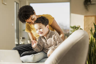 Smiling boy waving hand to video call on digital tablet while sitting by woman at home - UUF22650