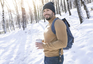 Lächelnder Mann träumt, während er eine Kaffeetasse im Schnee hält - JCCMF00831