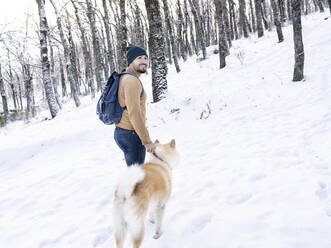 Smiling young man looking away while standing with akita dog in snow covered land - JCCMF00833