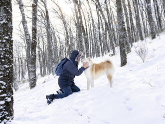 Mid adult man stroking dog while kneeling on snow land in forest - JCCMF00834