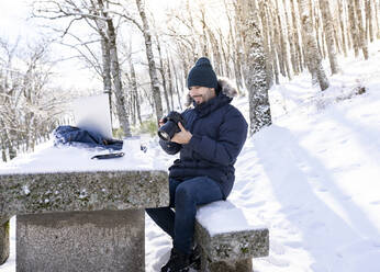 Lächelnder Fotograf mit Kamera auf einer Felsbank im Wald sitzend bei Schneewetter - JCCMF00836