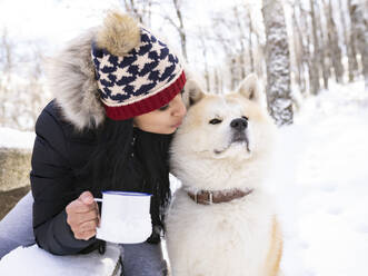 Mittlere erwachsene Frau mit Kaffeetasse küsst Hund, während sie auf einer Bank im Schnee sitzt - JCCMF00844