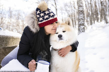 Frau mit geschlossenen Augen umarmt Akita-Hund und hält Kaffeetasse im Winter - JCCMF00845