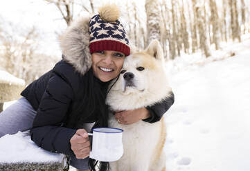 Glückliche Frau mit Kaffeetasse, die ihren Hund im Schnee umarmt - JCCMF00847