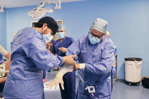 Assistentin, die dem Arzt hilft, einen Schutzhandschuh zu tragen, während er im Operationssaal steht, lizenzfreies Stockfoto