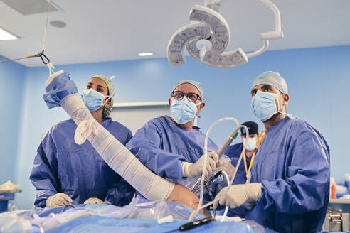 Ärzte mit Gesichtsmaske, die eine Operation mit Endoskopiegeräten durchführen, während sie im Operationssaal stehen - SASF00154