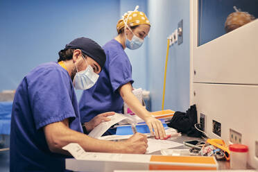 Assistentin mit Gesichtsmaske schreibt in Papier, während sie bei einer Krankenschwester im Krankenhaus arbeitet - SASF00168