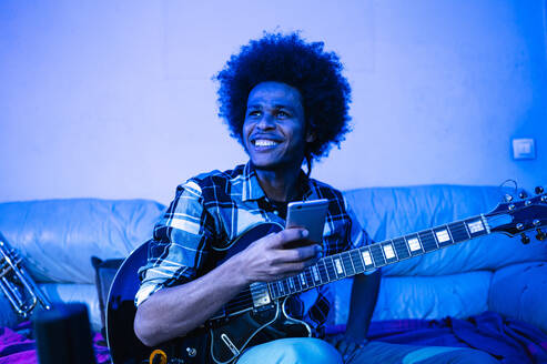 Glücklicher Afro-Mann mit Gitarre, der wegschaut, während er ein Mobiltelefon in einem blauen Raum hält - JCMF01797