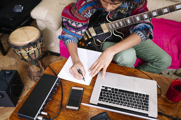 Junger Mann mit Gitarre schreibt Notizen auf einem Buch am Laptop im Wohnzimmer - JCMF01805