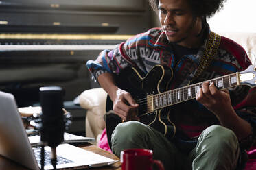 Afro young man learning to play guitar through laptop in living room - JCMF01812