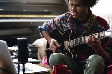 Ein junger Afroamerikaner lernt im Wohnzimmer über einen Laptop Gitarre spielen - JCMF01812