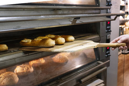 Male chef taking out freshly baked bread with pizza peel from oven at bakery - JCMF01819