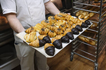 Männlicher Koch hält ein mit Croissants gefülltes Backblech in einer Bäckereiküche - JCMF01821