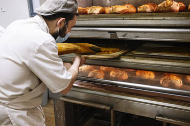 Männlicher Bäcker mit Pizzaschieber bei der Kontrolle von Brot im Ofen in einer Bäckerei während COVID-19 - JCMF01824