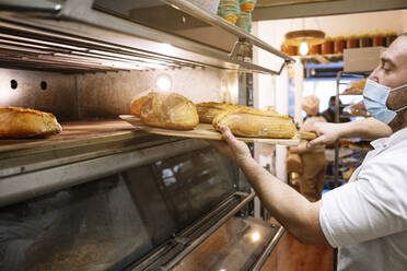 Männlicher Koch mit Gesichtsschutz, der in einer Bäckereiküche frisches Brot aus dem Ofen nimmt - JCMF01826