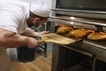 Männlicher Bäcker nimmt Brote aus dem Ofen in der Küche einer Bäckerei - JCMF01831