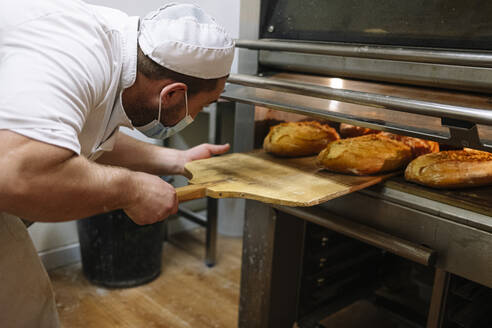 Male baker removing breads from oven in kitchen at bakery
 - JCMF01831
