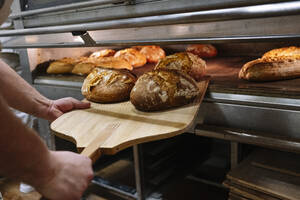 Männlicher Koch mit Pizzaschieber, der in einer Bäckerei das Brot aus dem Ofen nimmt - JCMF01832