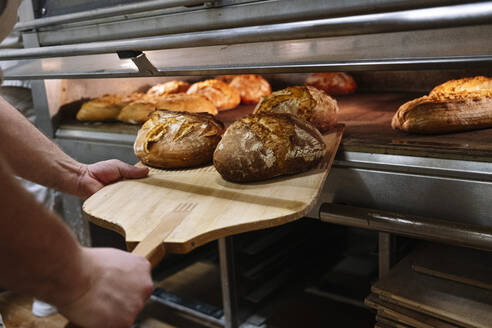 Male chef with pizza peel removing baked bread from oven at bakery - JCMF01832