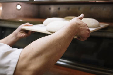 Männlicher Bäcker, der den Brotteig in einer Bäckerei im Ofen hält - JCMF01836