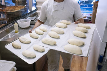 Männlicher Bäcker geht mit einem Backblech, das mit Mehlteig gefüllt ist, in der Küche einer Bäckerei - JCMF01837