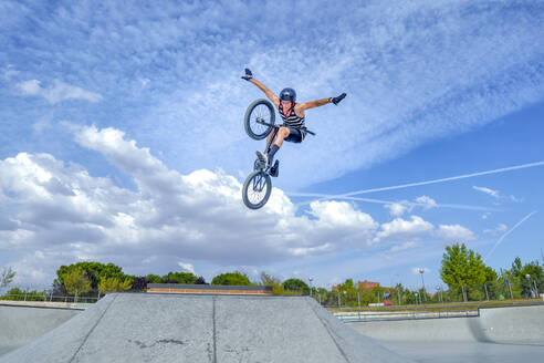 Carefree man with arms outstretched jumping with bicycle against blue sky - GGGF00787