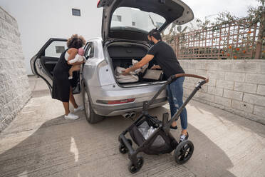 Rückenansicht eines ethnischen Mannes, der Gepäck und Kinderwagen in den Kofferraum legt, und einer afroamerikanischen Mutter mit Kleinkind, die ins Auto steigt, während sie sich auf die Reise vorbereitet - ADSF19806