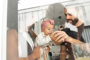 Low angle through window of multiracial couple giving juice for cute baby while sitting together at table in cafe - ADSF19808