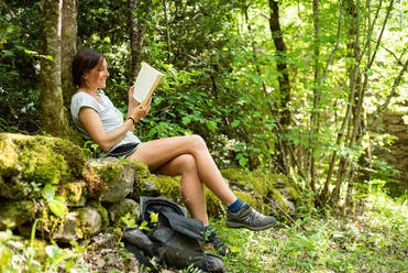 Seitenansicht einer konzentrierten Frau, die auf moosbewachsenen Steinen im Wald sitzt und eine interessante Geschichte in einem Buch genießt, während sie Zeit in der Natur verbringt - ADSF19811