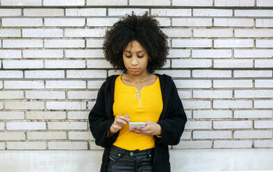 Beautiful woman with afro hair using smart phone while standing against brick wall - MGOF04628