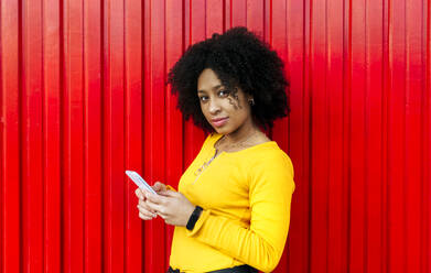 Selbstbewusste schöne Frau mit Afro-Haar, die ein Mobiltelefon vor einer roten Wand benutzt - MGOF04634