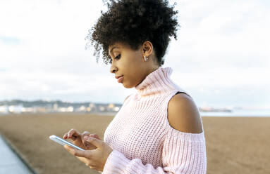 Schöne Frau mit lockigem Haar, die ein Mobiltelefon benutzt, während sie vor einem klaren Himmel sitzt - MGOF04642