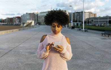 Junge Frau mit Afro-Haar, die ein Mobiltelefon benutzt, während sie auf der Straße steht - MGOF04646