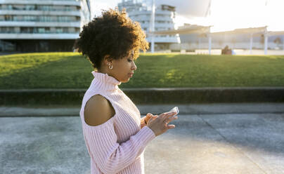 Woman with afro hair using mobile phone while standing on street in city - MGOF04647