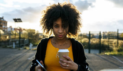 Junge Frau mit Afro-Haar hält Kaffee und Smartphone gegen den Himmel - MGOF04649