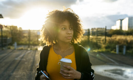 Junge Frau mit Afro-Haar hält Kaffee und Smartphone und schaut in den Himmel - MGOF04650