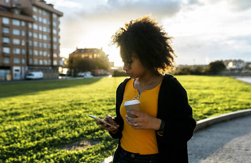 Junge Frau hält Kaffee und benutzt ein Mobiltelefon, während sie auf einem Fußweg gegen den Himmel läuft - MGOF04652