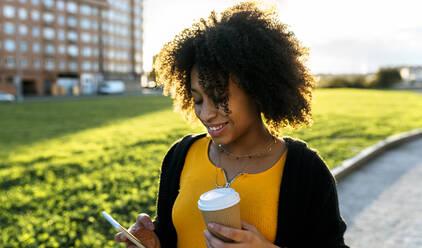 Lächelnde junge Frau mit lockigem Haar, die einen Kaffee in der Hand hält, während sie ein Mobiltelefon im Freien benutzt - MGOF04653