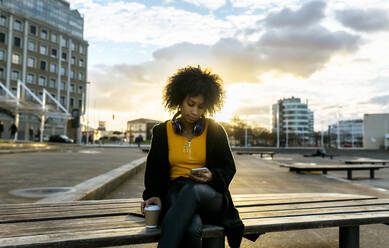 Junge Frau mit Afro-Haar, die ein Mobiltelefon benutzt, während sie auf einer Bank gegen den Himmel in der Stadt sitzt - MGOF04655