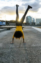 Junge Frau mit Afro-Haar macht Handstand auf der Straße gegen den Himmel bei Sonnenuntergang - MGOF04659