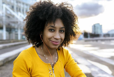 Nahaufnahme einer lächelnden Frau mit Afro-Haar, die über Kopfhörer Musik hört, gegen den Himmel - MGOF04661