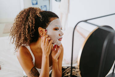 Young female with curly hair in mirror applying skincare moisturizing cloth mask on face during home beauty procedure - ADSF19829