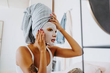 Young female in mirror applying skincare moisturizing cloth mask on face during home beauty procedure - ADSF19830
