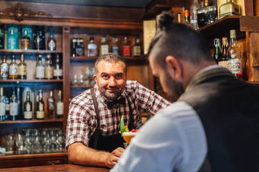 Barkeeper mittleren Alters schaut in die Kamera und gibt einem nicht erkennbaren eleganten bärtigen Mann einen Cocktail mit Himbeeren, während er in einer Bar arbeitet - ADSF19845