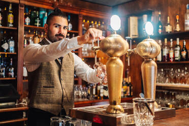 Konzentrierter erwachsener bärtiger Barkeeper zapft Bier in ein Glas, während er am Tresen in einer Kneipe arbeitet - ADSF19849