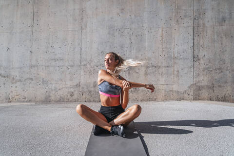 Slim female athlete with eyes closed in sportswear sitting on mat and stretching arms during workout in city stock photo