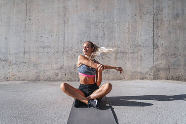 Slim female athlete with eyes closed in sportswear sitting on mat and stretching arms during workout in city - ADSF19859