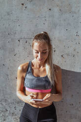 Cheerful female athlete in activewear leaning on wall and browsing mobile phone while resting during workout in city - ADSF19863