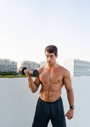 Athletic muscular shirtless guy doing fitness exercise with dumbbell during intense workout in city - ADSF19865