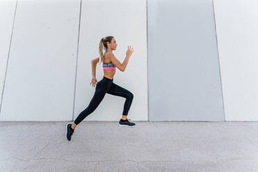 Side view of young fit female athlete in sportswear jogging during workout on city street and looking forward - ADSF19867